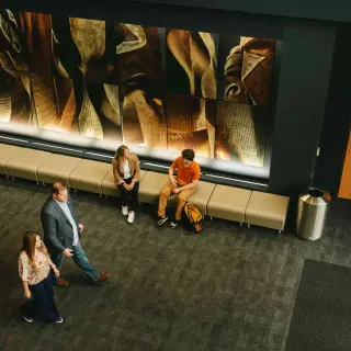 An overhead view of a young man in an orange polo talking to a woman in a blazer. They sit on cushioned benches along a wall covered with abstract artistic designs. Another man and woman walk past on the carpeted hallway in front of them.