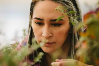 A young woman with silver-streaked dark hair stares intently at something out of frame. She looks focused and determined. She is surrounded by softly out-of-focus plants.