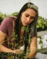 A young woman with long, dark, silver-streaked hair focuses intently while working on a project, a pen and notebook visible in her hands. She wears a burgundy and white-striped, short-sleeved shirt and is surrounded by plants in a greenhouse.