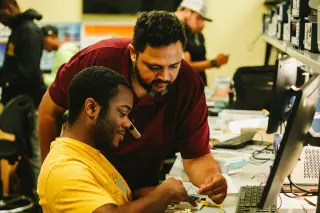 A man in a yellow shirt sits at a desk with a computer in front of him. He is working to rewire a piece of hardware as another man in a red polo leans over him to assist.
