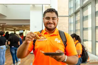 A young man in a bright orange polo shirt with the SHSU logo smiles at the camera while holding a slice of pizza in one hand and a black plastic plate in the other. He's wearing a grey backpack and is inside the Lowman Student Center; the nearby windows let in plenty of sunlight, while other students mingle in the background. 