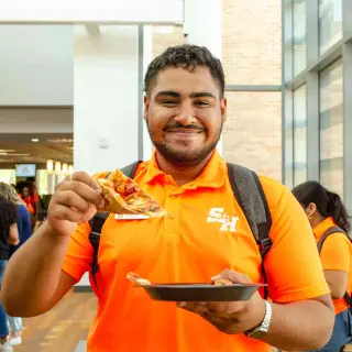 A young man in a bright orange polo shirt with the SHSU logo smiles at the camera while holding a slice of pizza in one hand and a black plastic plate in the other. He's wearing a grey backpack and is inside the Lowman Student Center; the nearby windows let in plenty of sunlight, while other students mingle in the background.