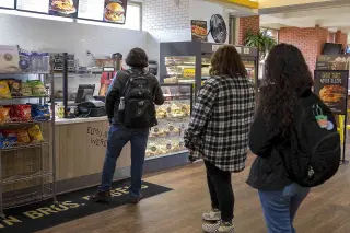 Three students stand in line at a campus food counter; the student at the front is placing their order while the other two students wait their turn. On the wall is the restaurant’s menu; there are wire shelves with various potato chip bags available for sale.