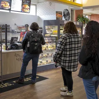 Three students stand in line at a campus food counter; the student at the front is placing their order while the other two students wait their turn. On the wall is the restaurant’s menu; there are wire shelves with various potato chip bags available for sale.