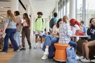 A wide shot of the bustling Lowman Student Center filled with students walking, talking, eating, and sitting together. Sunlight spills in through the floor-to-ceiling windows, while SHSU orange provides accent coloring on the carpet and the chairs. It feels lively and busy. 