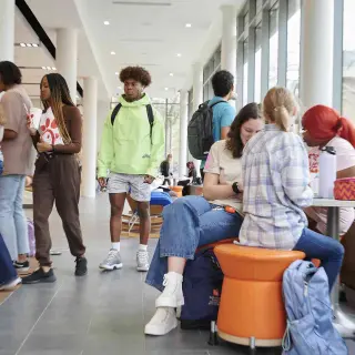 A wide shot of the bustling Lowman Student Center filled with students walking, talking, eating, and sitting together. Sunlight spills in through the floor-to-ceiling windows, while SHSU orange provides accent coloring on the carpet and the chairs. It feels lively and busy.