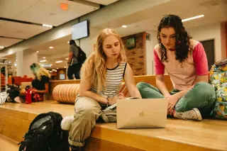 Two students, both young women, study together in the Lowman Student Center. One student with long blonde hair wearing a striped shirt is writing in a notebook, while another with curly dark hair in a pink shirt is looking at a laptop. They're sitting on large wooden step-style seating, with backpacks nearby and other students visible in the background.