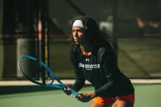 A tennis player on a court holds her tennis racket in a ready position; she is wearing a black Sam Houston Tennis shirt, orange shorts, and a white headband. She appears focused, with an intense look on her face.