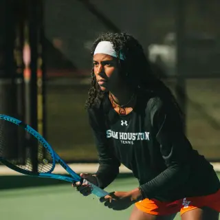 A tennis player on a court holds her tennis racket in a ready position; she is wearing a black Sam Houston Tennis shirt, orange shorts, and a white headband. She appears focused, with an intense look on her face.