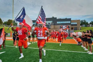 Two SHSU football players in orange uniforms, numbered 96 and 21, run onto the football field carrying the Texas and American flags. The rest of the football team follows behind them; cheerleaders, dressed in black uniforms and holding alternating orange and white pom-poms, line the field to cheer the players on.