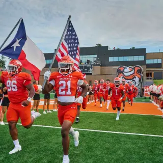 Two SHSU football players in orange uniforms, numbered 96 and 21, run onto the football field carrying the Texas and American flags. The rest of the football team follows behind them; cheerleaders, dressed in black uniforms and holding alternating orange and white pom-poms, line the field to cheer the players on.