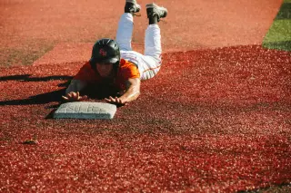 A baseball player slides headfirst into base on a red dirt infield. Wearing an orange jersey, white pants, and black helmet, the player reaches for the plate as he slides, a look of determination on his face.