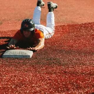 A baseball player slides headfirst into base on a red dirt infield. Wearing an orange jersey, white pants, and black helmet, the player reaches for the plate as he slides, a look of determination on his face.