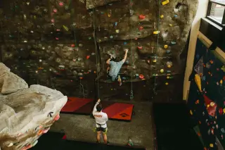 A wide angle shot of an indoor rock-climbing wall. A climber in a light blue shirt scales the wall; a staff member in a white shirt belays him. Red crash pads are visible on the floor, and there's a mixture of climbing surfaces off to the sides of the frame—to the left, an artificial rock formation, and on the right, traditional climbing walls with various routes marked by different colored holds.