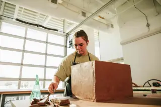 A young woman wearing an apron assembles a box made of clay in a classroom with a wall of windows behind her.