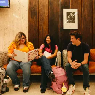 Three students sit across two loveseat couches in the Newton Gresham Library; there is a wood-paneled wall behind them. One student, a young woman, wears a yellow shirt and glasses while holding out at a white binder for the other students to look at with her. The other two students are a young woman wearing a pink shirt and holding a book, and a young man, wearing a black shirt. They are all smiling and talking with each other.