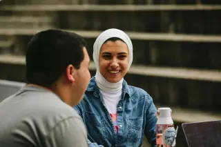 In an outside amphitheater seating area, a young woman, wearing a white hijab and a denim button-up shirt, smiles while engaging in conversation with a young man whose face is turned away from the camera. She holds a pink water bottle and is looking at the young man’s laptop.