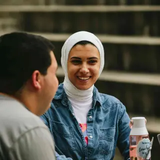 In an outside amphitheater seating area, a young woman, wearing a white hijab and a denim button-up shirt, smiles while engaging in conversation with a young man whose face is turned away from the camera. She holds a pink water bottle and is looking at the young man’s laptop.