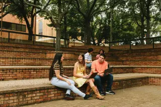 Five students gather in an outdoor amphitheater seating area. In the foreground, three students, two women and one man, sit together talking—one woman wears a black and white striped shirt, the other is in a yellow dress with cowboy boots, and a man wears a coral-colored shirt; the latter two students have their laptops open. Two more students can be seen sitting higher up on the steps in the background, also conversing. Trees dot the steps around them.