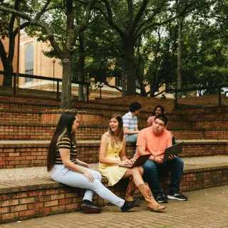 Five students gather in an outdoor amphitheater seating area. In the foreground, three students, two women and one man, sit together talking—one woman wears a black and white striped shirt, the other is in a yellow dress with cowboy boots, and a man wears a coral-colored shirt; the latter two students have their laptops open. Two more students can be seen sitting higher up on the steps in the background, also conversing. Trees dot the steps around them.