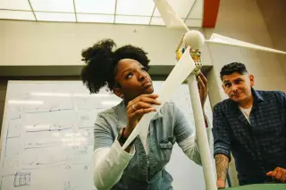 A student examines a model wind turbine in a large classroom with a whiteboard behind her, as her professor looks on.