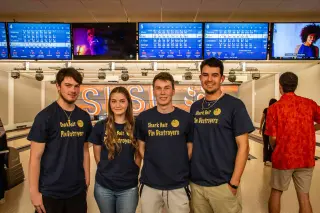 Four students pose together in Sam Houston State University’s bowling alley. They all wear black t-shirts with the text “Shark Bait Pin Destroyers” a bowling ball on them. Screens displaying bowling scores show above their heads.