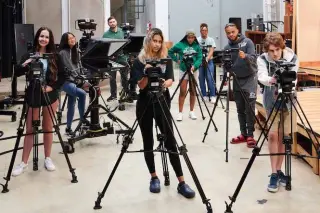 A group of students in a media production studio, each operating their own professional video cameras mounted on tripods. The setting includes various production equipment, such as lights, monitors, and a short wooden stage. Some students smile and engage with the photographer’s camera, while others look down, focusing on their video cameras.