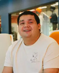 A young man, sitting in an orange-colored chair, smiles, looking up and away from the camera. The young man is brunette and wears a white shirt.
