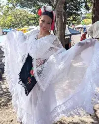 A young woman wearing traditional Mexican folkloric dance attire smiles at the camera. She wears a white lace shawl over a white blouse and black skirt with floral embroidery; her arms are raised shoulder-height, to better show her attire. She has a decorative red and white hair accessory with flowers and ribbons; she stands under a tree, shaded from the sun.