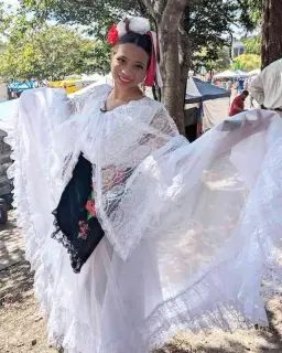 A young woman wearing traditional Mexican folkloric dance attire smiles at the camera. She wears a white lace shawl over a white blouse and black skirt with floral embroidery; her arms are raised shoulder-height, to better show her attire. She has a decorative red and white hair accessory with flowers and ribbons; she stands under a tree, shaded from the sun.