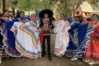A group of performers in traditional Mexican folkloric attire poses together outdoors. The women wear colorful, flowing dresses adorned with rainbow-patterned stripes and lace, while the man in the center wears a black charro suit with intricate embroidery and a large sombrero. The background features trees and festive decorations, making it feel celebratory.