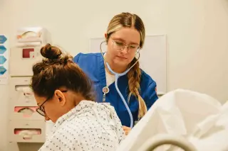 A nursing student wearing blue scrubs and a stethoscope presses the other end of the stethoscope against the back of a young woman wearing a hospital gown and glasses. They are in a hospital room.