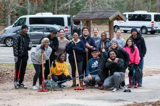A group of students dressed in chilly weather clothing pose and smile for the camera in a parking lot. Some of them are holding various gardening tools, including rakes, shears, and gardening hoes. The students have just finished some outdoor community service. 