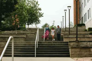 Three students walk down a wide staircase outside near one of the residence halls. The stairs have white handrails and are flanked by stone walls and decorative lampposts. One student, a young woman, is wearing a flowing pink skirt, denim button-up shirt, and white hijab; another young woman wears a shirt and shorts; the third, a young man, is in dark athleisure wear. Green trees line the top of the staircase, and a white building is visible on the right.