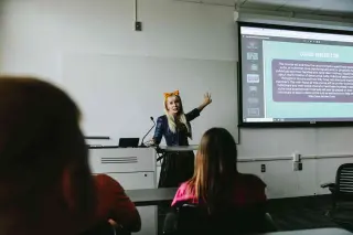 A criminal justice instructor in a dark dress and yellow bow in her blonde hair stands at the front of a classroom, gesturing with one hand raised while teaching. Behind her, a projected slide shows text about a course description related to the study of cults. Students can be seen from behind in the foreground, seated at desks. 