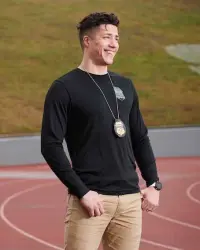 A smiling young man with short, taper-faded hair stands outdoors against a grassy background; he is looking off into the distance. He is wearing a black long-sleeved shirt, and a law enforcement badge hangs around his neck.