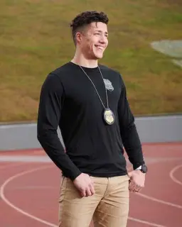 A smiling young man with short, taper-faded hair stands outdoors against a grassy background; he is looking off into the distance. He is wearing a black long-sleeved shirt, and a law enforcement badge hangs around his neck.