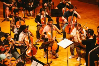 A group of young musicians rehearsing in an orchestra, photographed from an elevated angle. There are several string players, including multiple cellists and violinists, with their music stands and sheet music visible. The musicians are dressed casually for rehearsal. The lighting gives the scene a warm, golden tone.