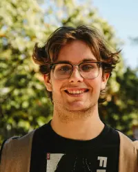 A young man wearing a black shirt with lettering, tan zip-up hoodie, and eyeglasses smiles at the camera. Sun-dappled trees are visible in the background.