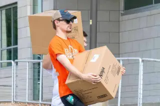 A young man wearing an orange SHSU shirt, baseball cap, and sunglasses carries a large cardboard moving box towards a residence hall; another man behind him also carries a cardboard box.