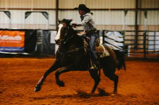 A SHSU student wearing a cowboy hat, a button-up shirt, and jeans rides her horse inside the rodeo arena. The horse, dark brown with a white blaze on its face, is galloping, and the student looks forward intently. The red dirt floor, banners, and fencing are visible in the background.