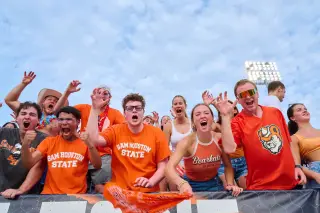A group of enthusiastic SHSU students cheer in the stands during an athletic game. They’re wearing SHSU-branded shirts while doing the “Eat ‘em up, ‘Kats!” hand motion—raising their right hands in a claw gesture.