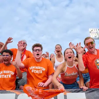 A group of enthusiastic SHSU students cheer in the stands during an athletic game. They’re wearing SHSU-branded shirts while doing the “Eat ‘em up, ‘Kats!” hand motion—raising their right hands in a claw gesture.