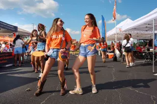 Two students, both young women, in orange SHSU shirts and denim shorts laugh together at a tailgating event. One wears cowboy boots while the other wears sneakers. Behind them are white tents with university organization banners, including "Business Administration" and "Student Government Association.” In the background, other students browse through the tents.