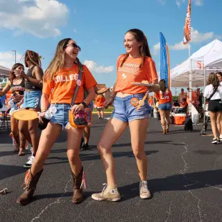 Two students, both young women, in orange SHSU shirts and denim shorts laugh together at a tailgating event. One wears cowboy boots while the other wears sneakers. Behind them are white tents with university organization banners, including "Business Administration" and "Student Government Association.” In the background, other students browse through the tents.