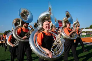 Members of the SHSU marching band's tuba section walking onto the football field, wearing orange and black uniforms. One student, in the center of the frame, smiles brightly at the camera while holding her sousaphone tuba. The other tuba players are visible in the background, their instruments gleaming in the sunlight.