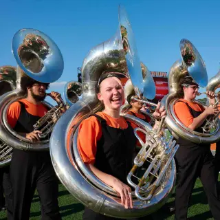 Members of the SHSU marching band's tuba section walking onto the football field, wearing orange and black uniforms. One student, in the center of the frame, smiles brightly at the camera while holding her sousaphone tuba. The other tuba players are visible in the background, their instruments gleaming in the sunlight.