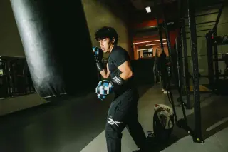 A young man in black workout clothing and boxing gloves trains with a punching bag in a dimly lit gym. He stands in a fighting stance, ready to make his next punch. Exercise equipment and weight racks are visible in the background.