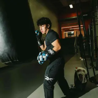 A young man in black workout clothing and boxing gloves trains with a punching bag in a dimly lit gym. He stands in a fighting stance, ready to make his next punch. Exercise equipment and weight racks are visible in the background.