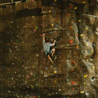 A young man wearing a gray shirt, black shorts, and climbing shoes scales an indoor rock-climbing wall. The wall is dotted with colorful hand- and footholds, secured with climbing ropes. The climber appears focused and mid-action, using their hands and feet to maintain grip on the vertical surface.